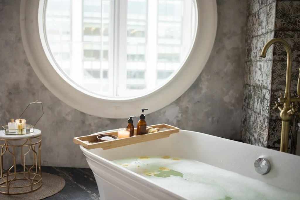 White freestanding bathtub beneath a round window with bath accessories and textured bathroom walls